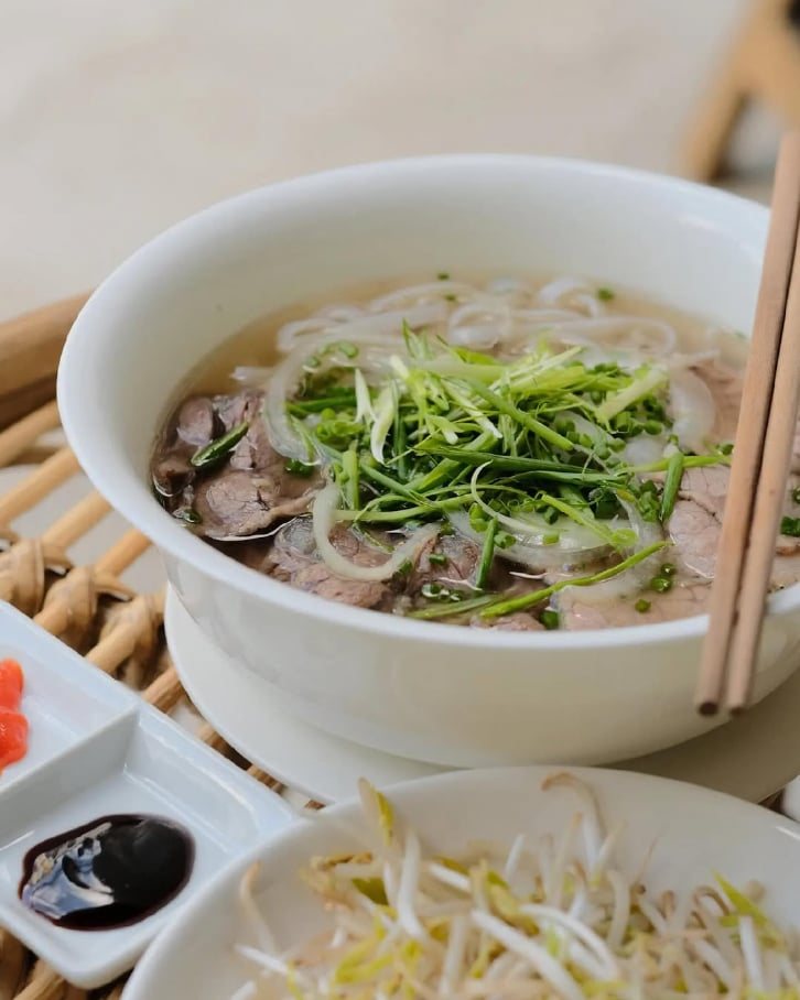 Bowl of beef phở topped with spring onion, with bean sprouts and sauces on the side