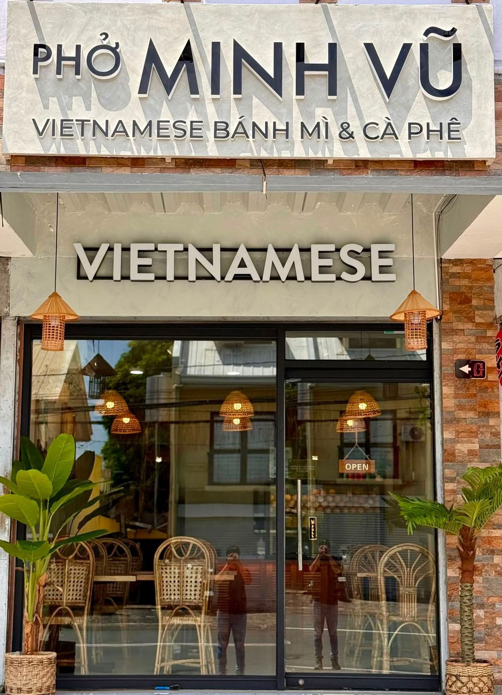 The Phở Minh Vũ BF Homes storefront in daylight, with its sign, glass door, rattan chairs and plants