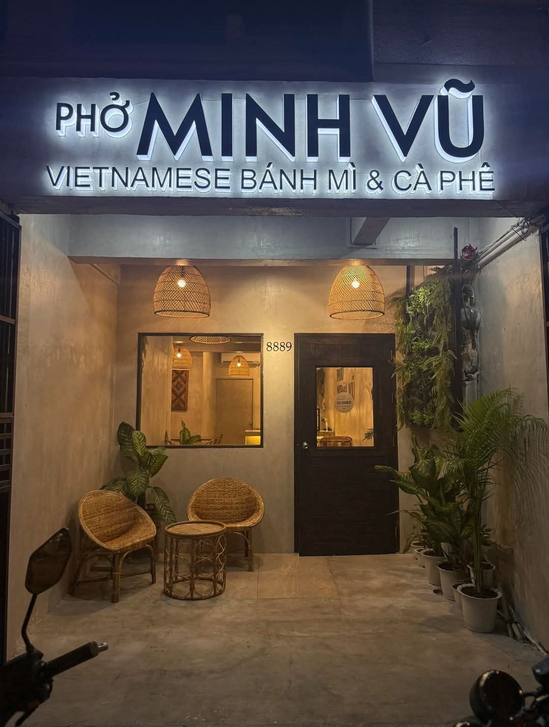 The Phở Minh Vũ Makati storefront at night, with its lit sign, rattan chairs, hanging lamps and plants by the door