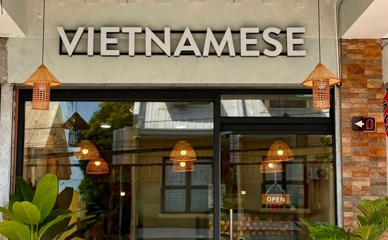 Front of the Phở Minh Vũ BF Homes branch with its Vietnamese sign, glass door, lanterns and rattan chairs